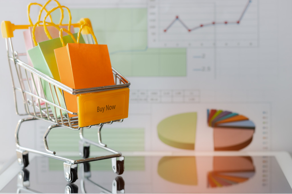 Colourful paper bags in yellow trolley on tablet with chart background. Consumers can buy products directly from seller over internet using web browser. Online shopping and e-commerce concept.