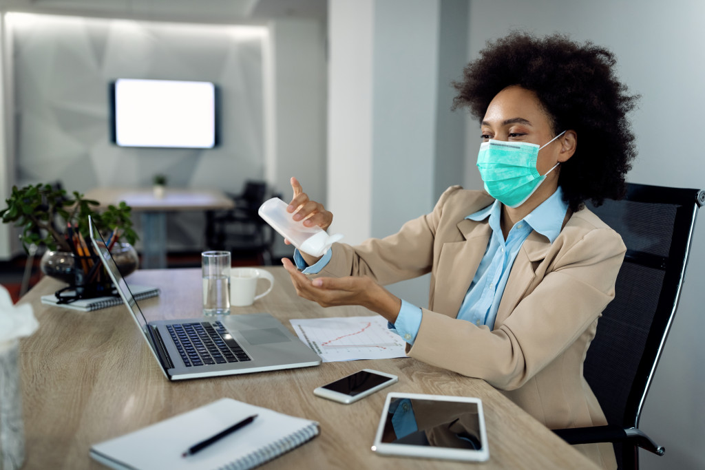 African American businesswoman using hands sanitizer and wearing face mask while working in the office during coronavirus pandemic.
